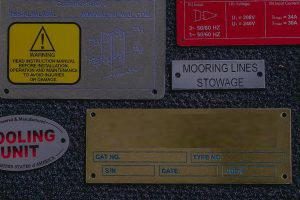 Assorted engraved and printed metal nameplates including safety labels, part ID tags, and brass data plates, produced in Bay Area California.