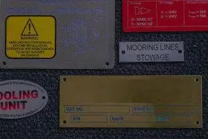 Assorted engraved and printed metal nameplates including safety labels, part ID tags, and brass data plates, produced in Bay Area California.